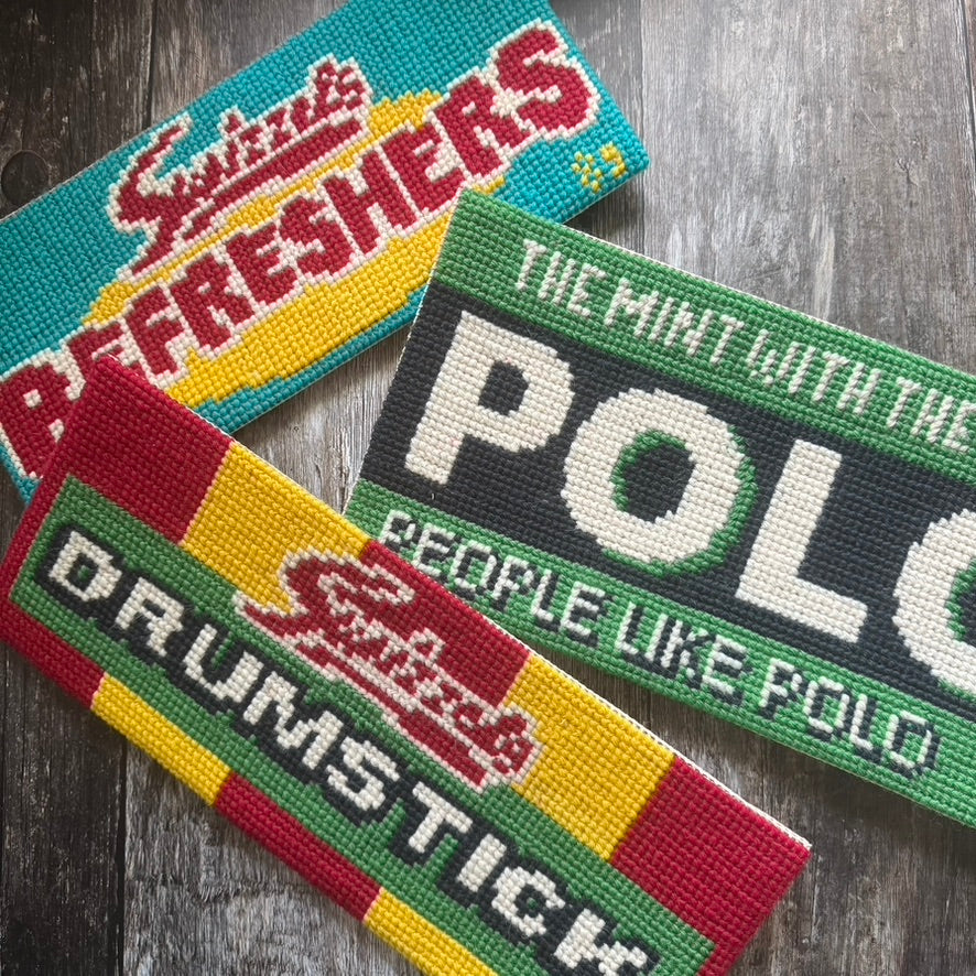Three colorful rectangular  cross stitch tuck shop kits by jacqui pearce on a wooden surface